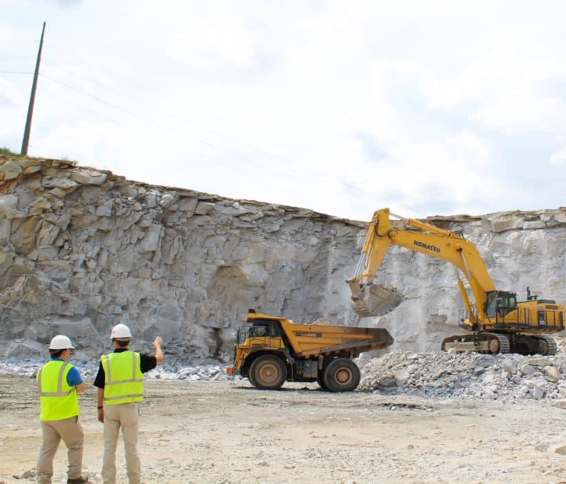 Two construction workers in safety vests and helmets observe an excavator loading rocks into a dump truck at a quarry. The backdrop features a rocky cliff and a cloudy sky.