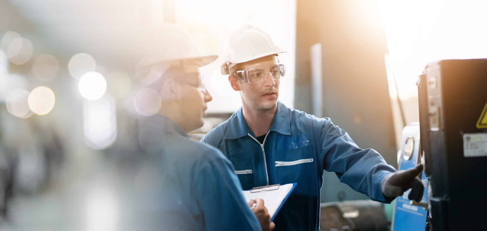 Two workers wearing safety helmets and goggles collaborate in an industrial setting. One holds a clipboard while the other operates machinery, with soft lighting suggesting a professional workplace environment.