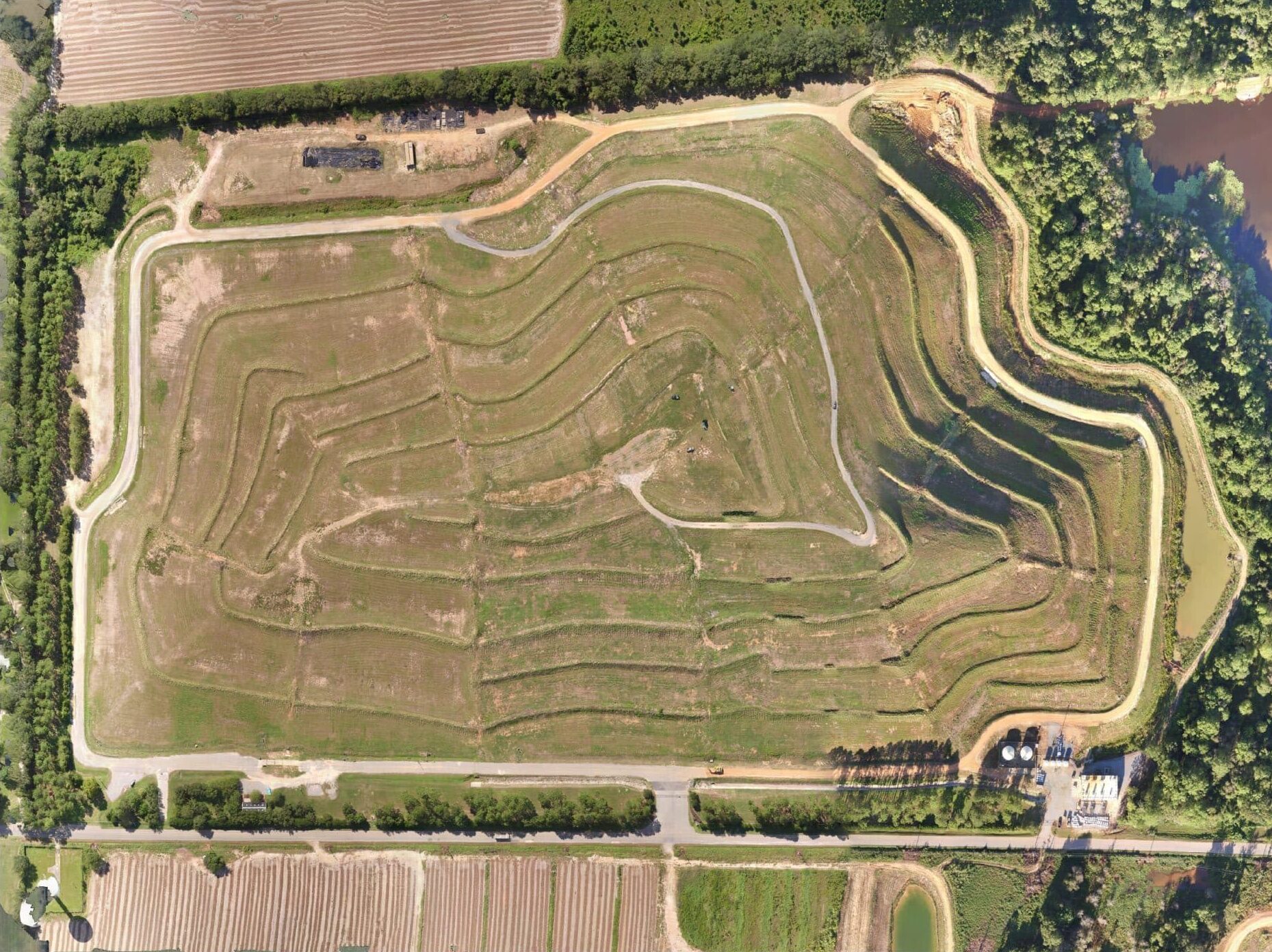 Aerial view of a motocross track with winding dirt paths and curves surrounded by greenery and fields. The track features multiple layers and intricate loops, indicating a course designed for high-speed racing and skillful navigation.