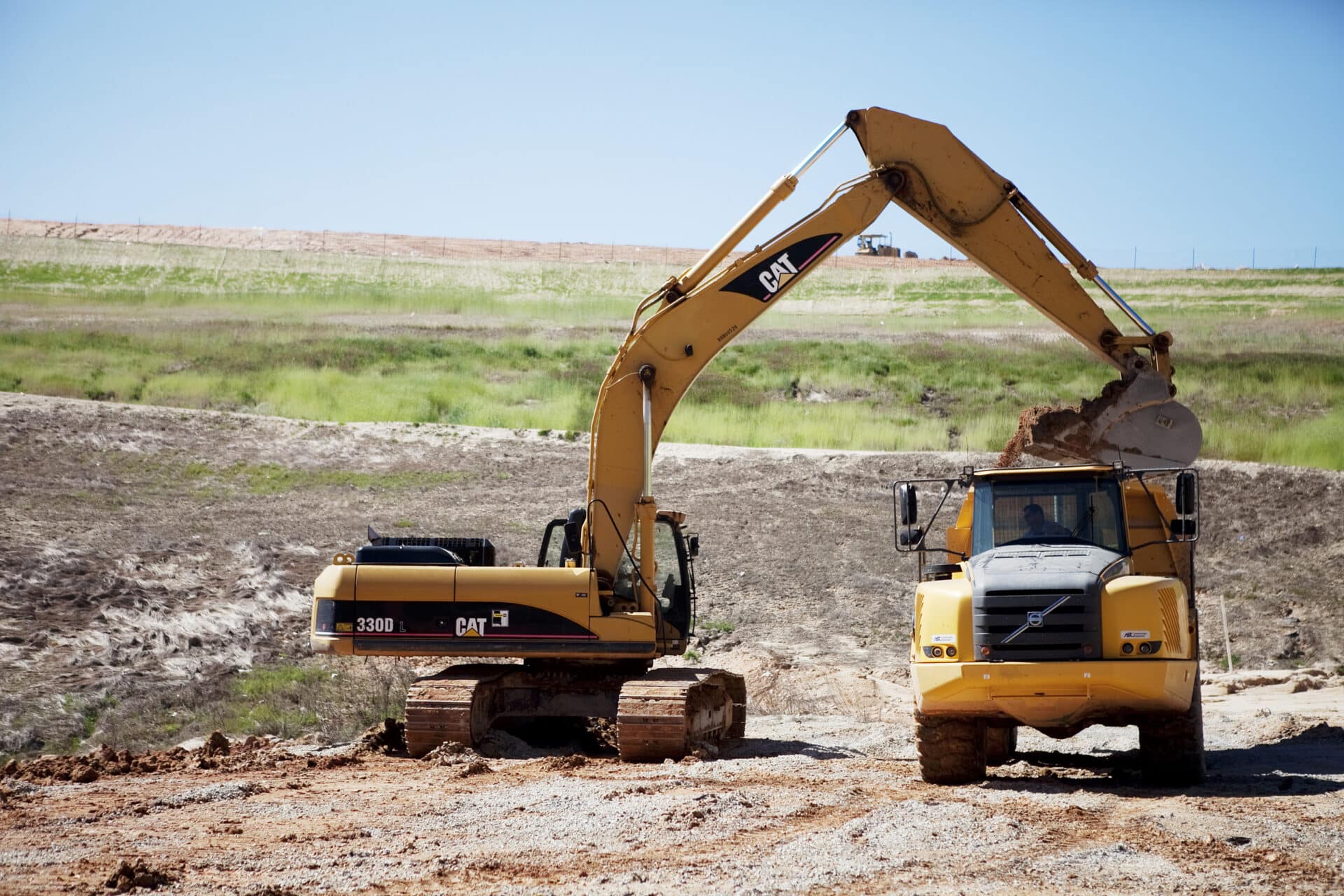 A yellow excavator loads dirt into a yellow dump truck at a construction site. The background features a grassy landscape and a clear blue sky.