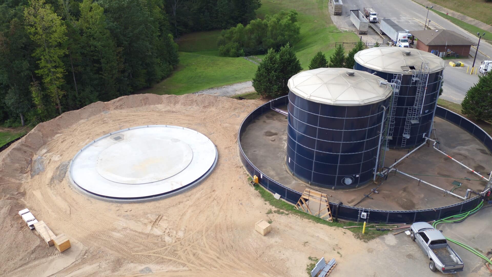 Aerial view of a construction site with two large cylindrical tanks, a circular concrete foundation, and construction materials. Surrounded by trees and a road in the background.