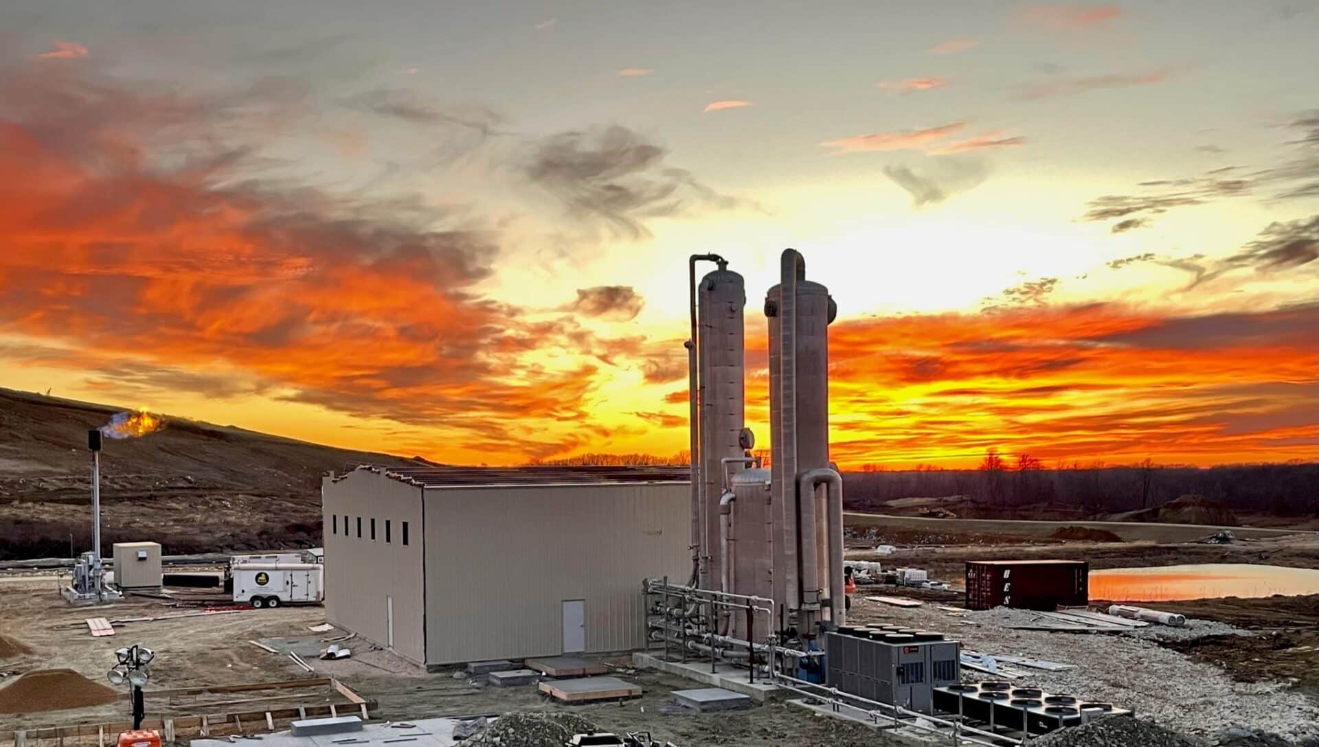 A factory with tall cylindrical structures is silhouetted against a vibrant sunset. The sky is filled with orange, yellow, and pink clouds. The surrounding area includes open land, machinery, and construction materials.