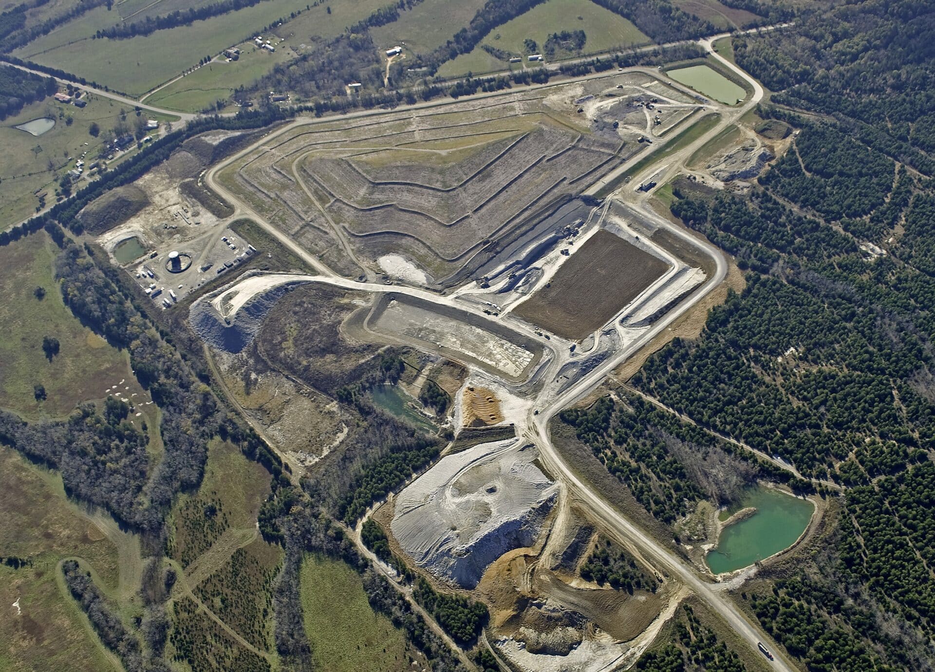 Aerial view of a large landfill site surrounded by fields and forested areas. The landfill has multiple tiered layers, enclosed roads, and a few small ponds. A network of roads connects different sections of the area.