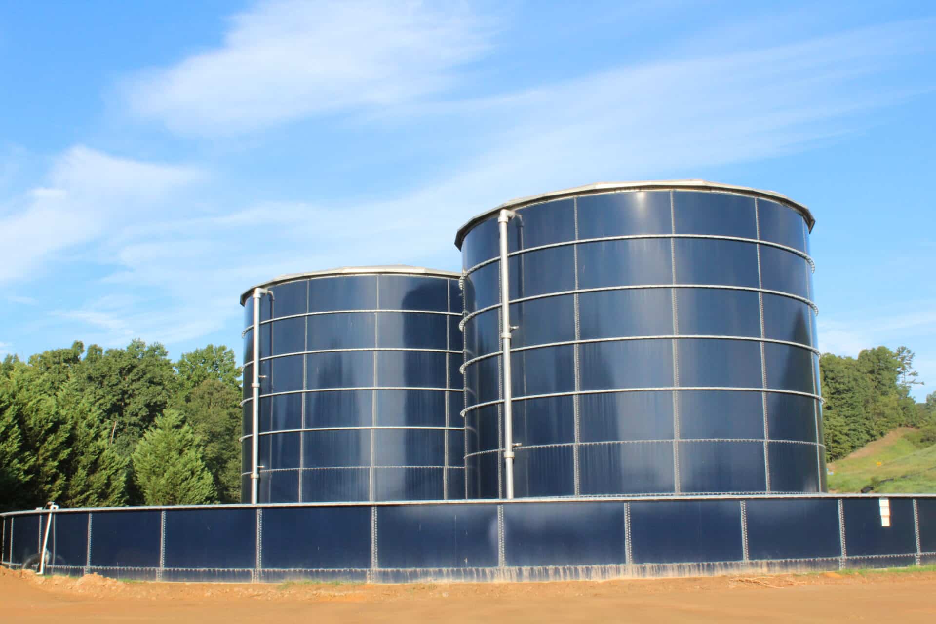 Two large, cylindrical blue water tanks are situated outdoors against a backdrop of green trees and a clear blue sky. The tanks are surrounded by a low fence, and the ground is bare and brown.