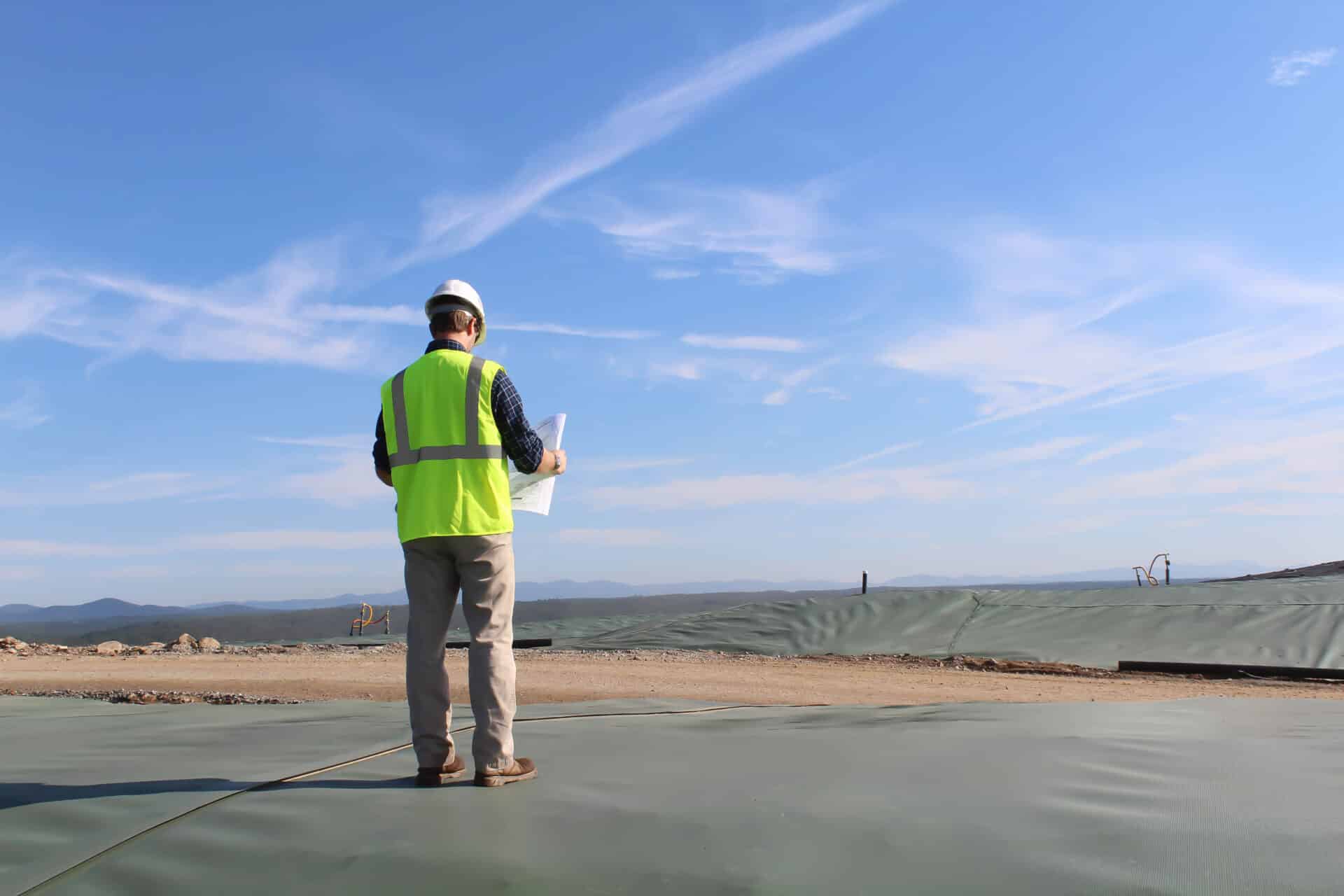 A construction worker in a yellow safety vest and hard hat stands on a large, covered area, holding documents. The sky is clear and blue, and the landscape is expansive with distant mountains.