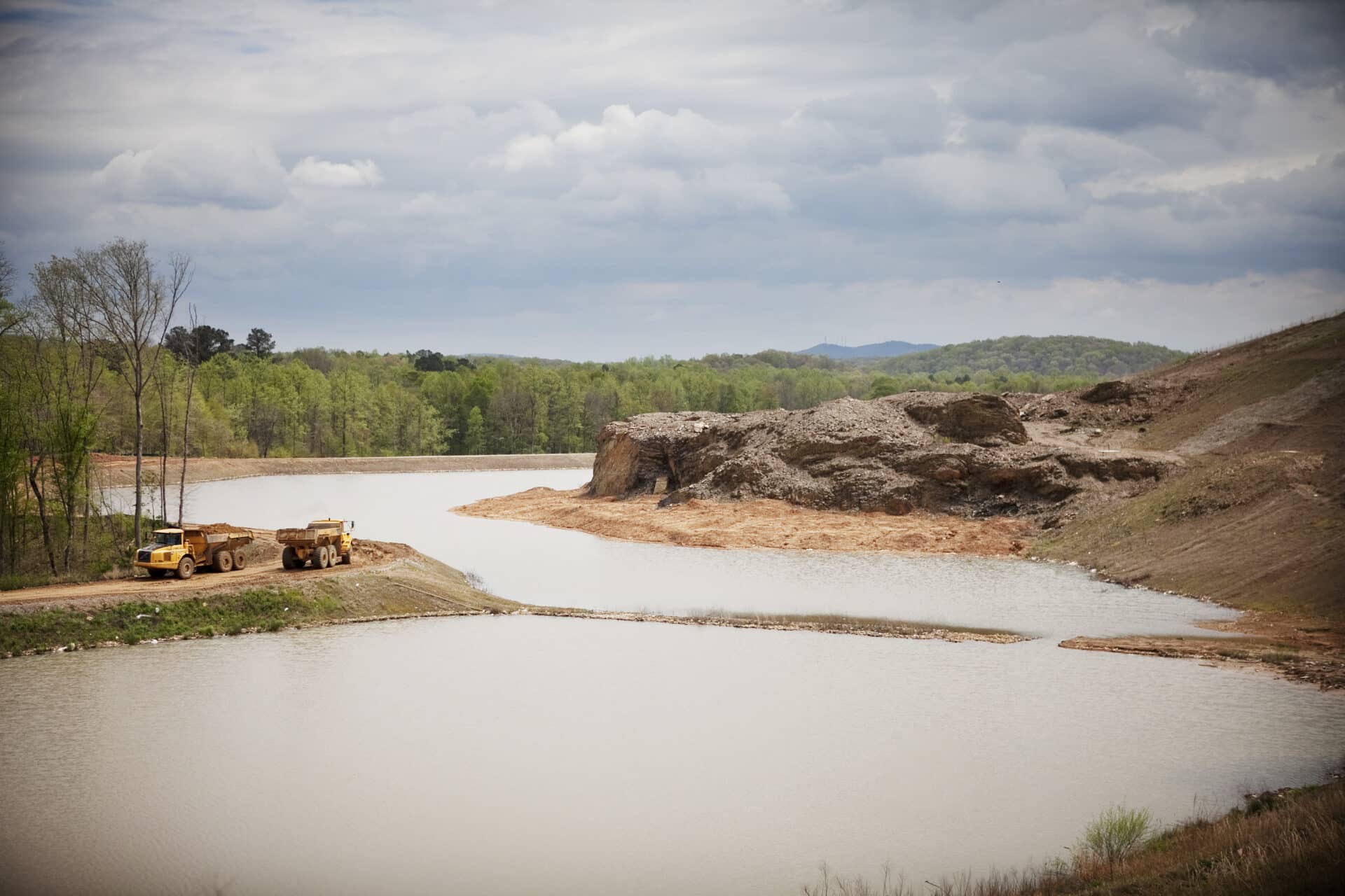 A scenic view of a quarry with a lake and surrounding greenery. Two large trucks are parked on a dirt road along the water's edge. The sky is cloudy, adding a dramatic effect to the landscape.