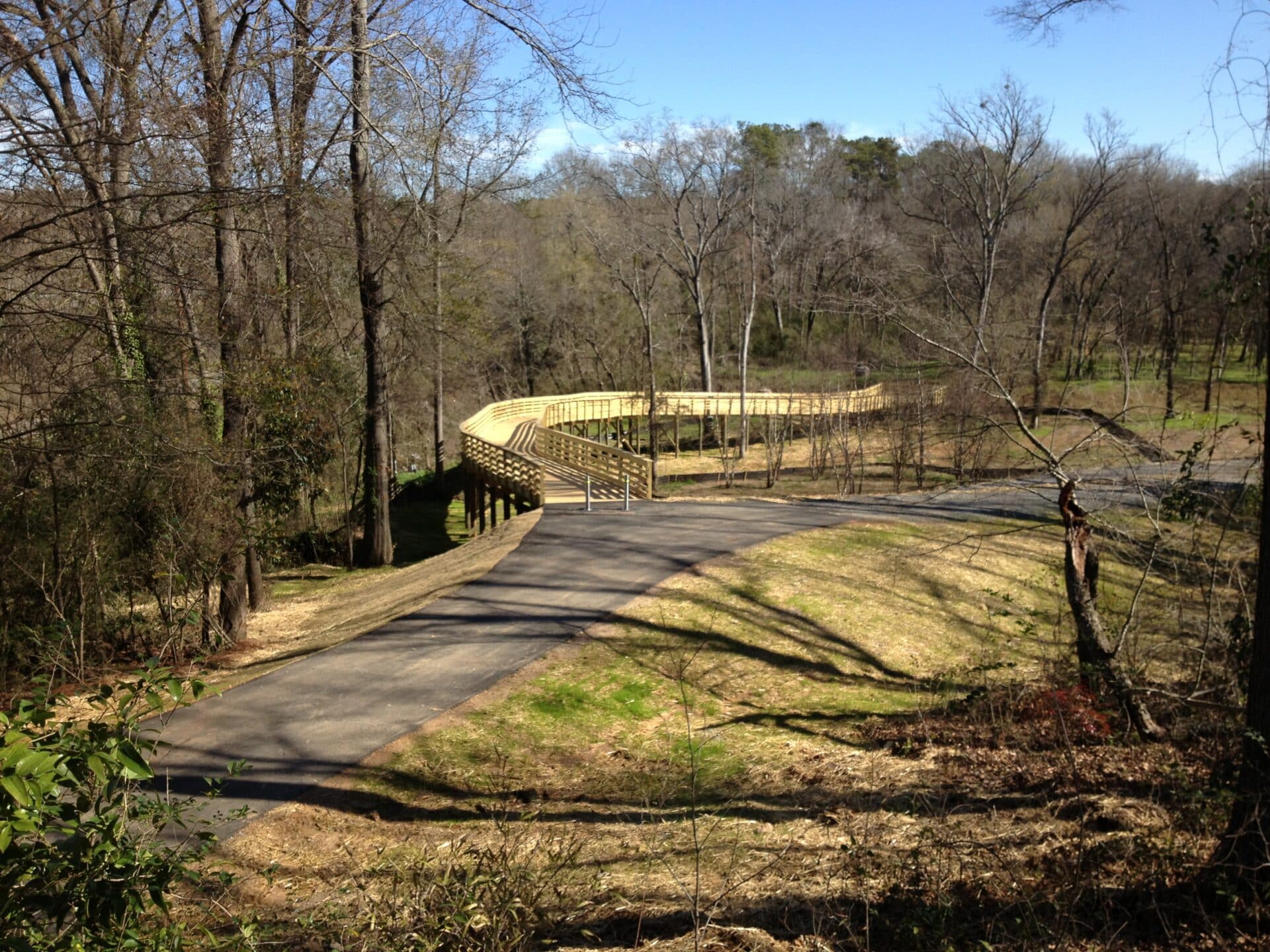 A winding wooden boardwalk curves through a forested area, surrounded by leafless trees and grassy patches. The sky is clear and blue, hinting at a sunny day.