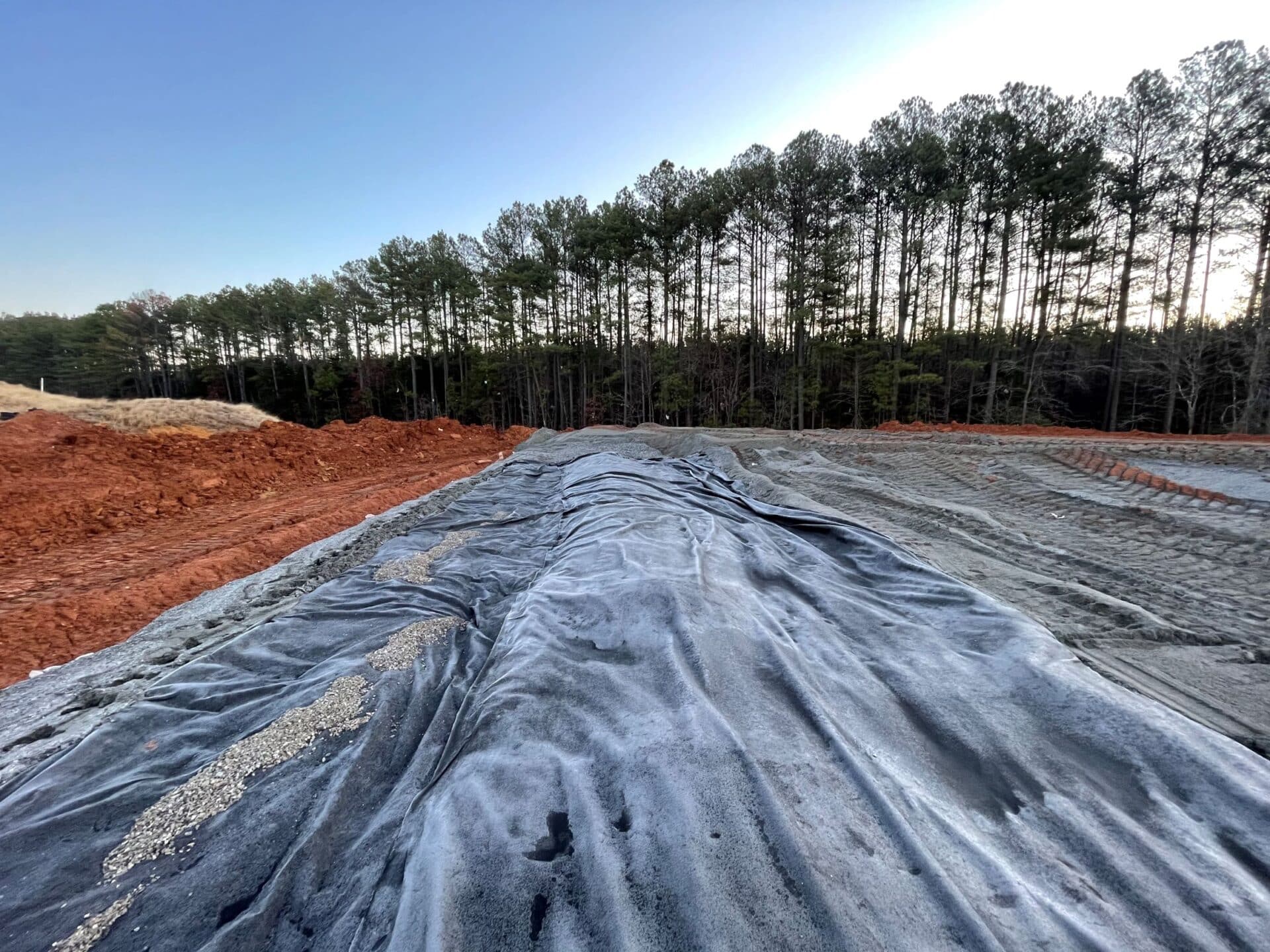 A landscape view of a construction site with red soil and a large black tarp covering the ground. In the background, there is a row of tall pine trees under a clear blue sky.