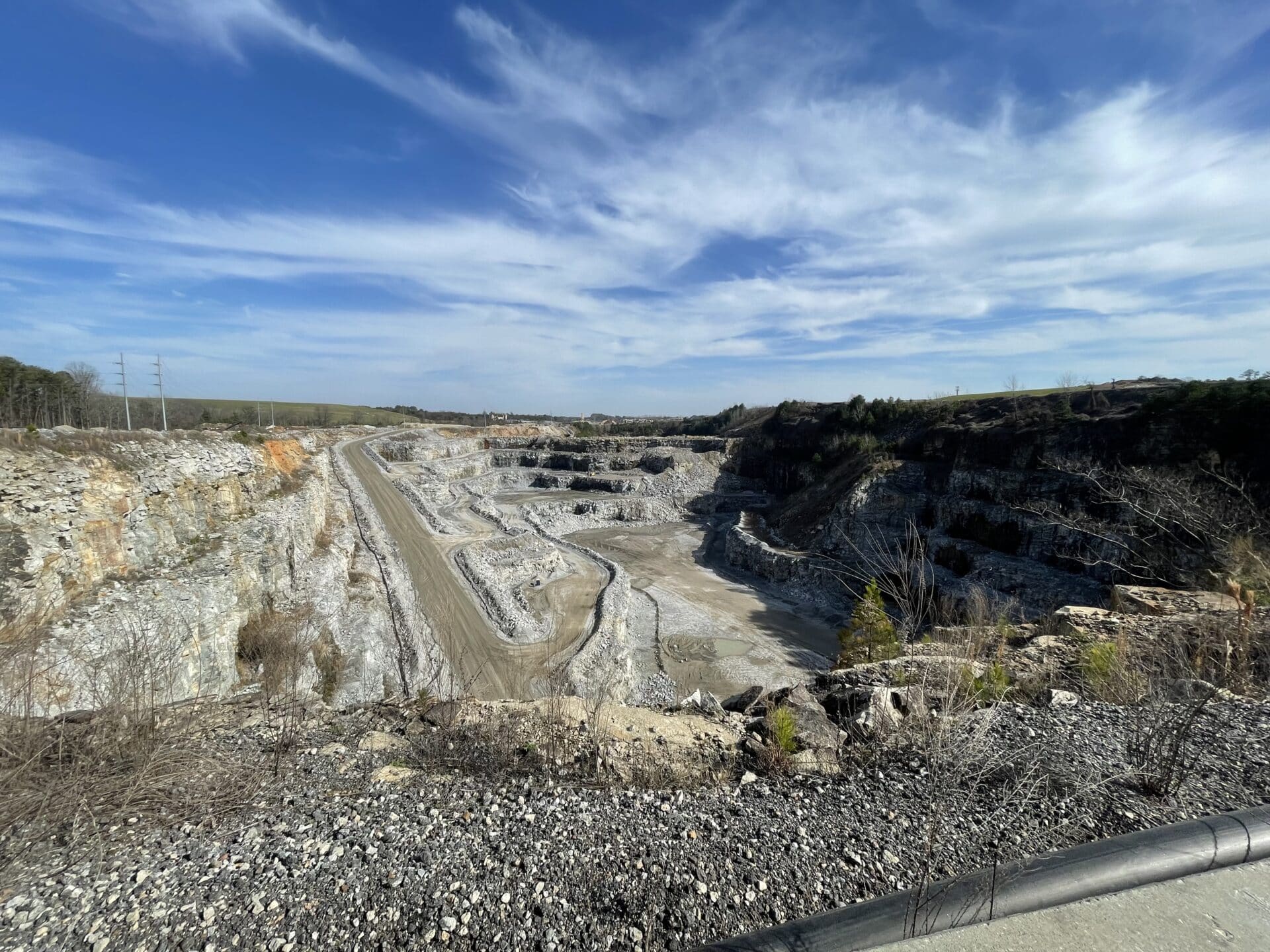 A panoramic view of a large open-pit mine with terraced rock layers. The sky above is partly cloudy and blue, lending a sense of vast openness. Sparse vegetation can be seen along the edges of the quarry.