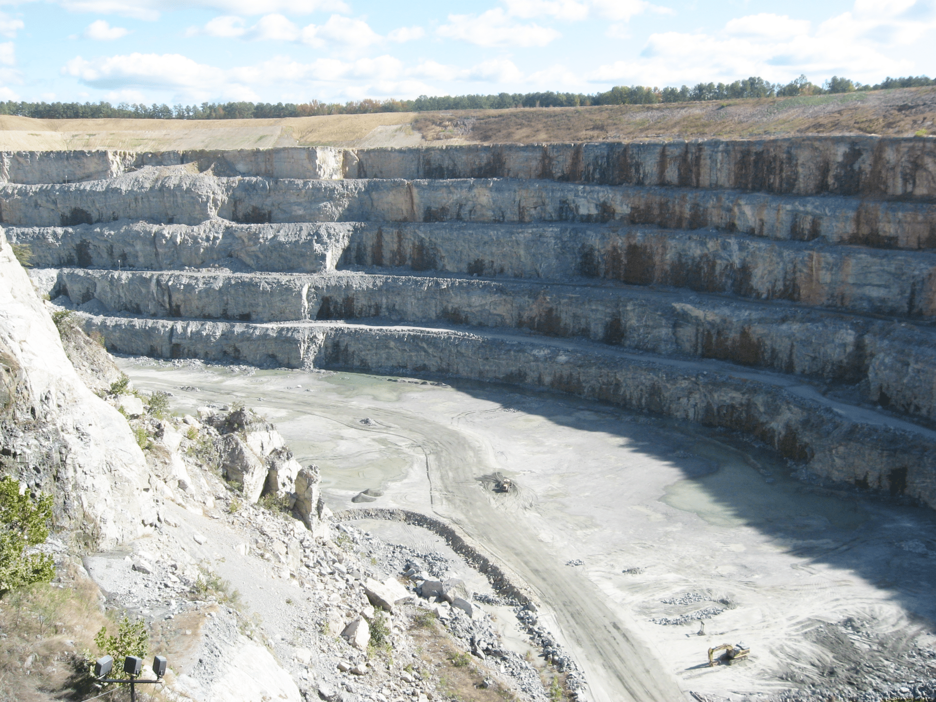 A large, multi-layered stone quarry with terraced levels. The excavation site is mostly barren with some patches of dirt and small pools of water at the bottom. Blue skies and trees are visible in the background.