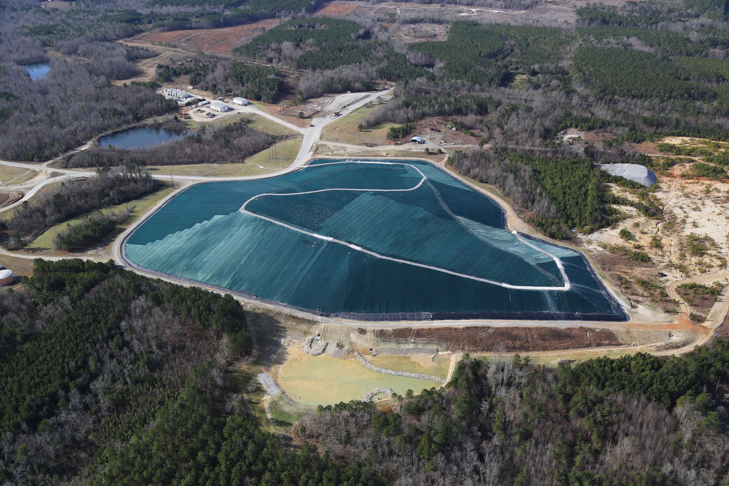 Aerial view of a landfill covered with a large blue tarp, surrounded by forested areas and dirt roads. Several small buildings and other structures are visible in the background. The landscape is a mix of trees and cleared land.