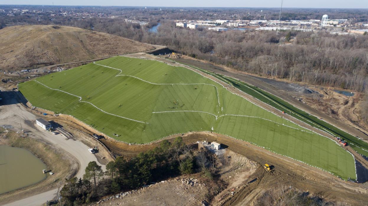 Aerial view of a large green landfill covered with synthetic turf, nestled in a rural area with trees and a dirt road. Construction equipment and materials are visible around the site. Houses and a town are in the distant background.