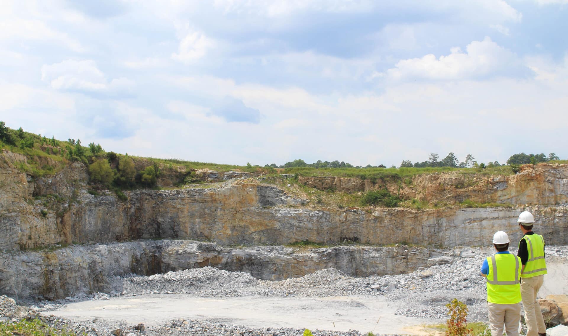 Two construction workers wearing safety gear stand at the edge of a large, rocky quarry. The sky is partly cloudy, and the surrounding terrain is green with distant trees.