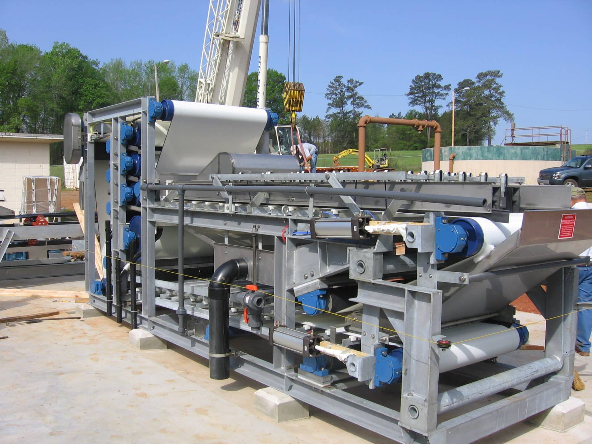 An industrial machine with conveyor belts and rollers, set up outdoors on a concrete surface. A crane and workers can be seen in the background, with trees and a clear blue sky above.