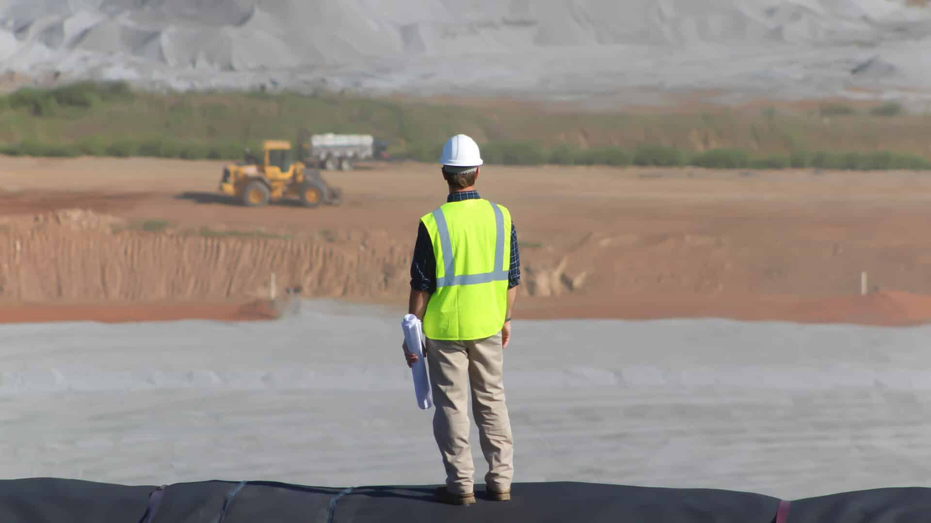 A construction worker wearing a white hard hat and neon yellow safety vest stands on a raised platform, overlooking a vast construction site with earth-moving equipment in the distance.