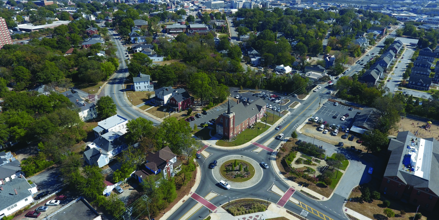 Aerial view of a small town with a roundabout at the center. Surrounding the roundabout are residential houses, a few larger buildings, and parking lots. The area is lush with greenery, and the town extends towards the horizon with more structures visible.