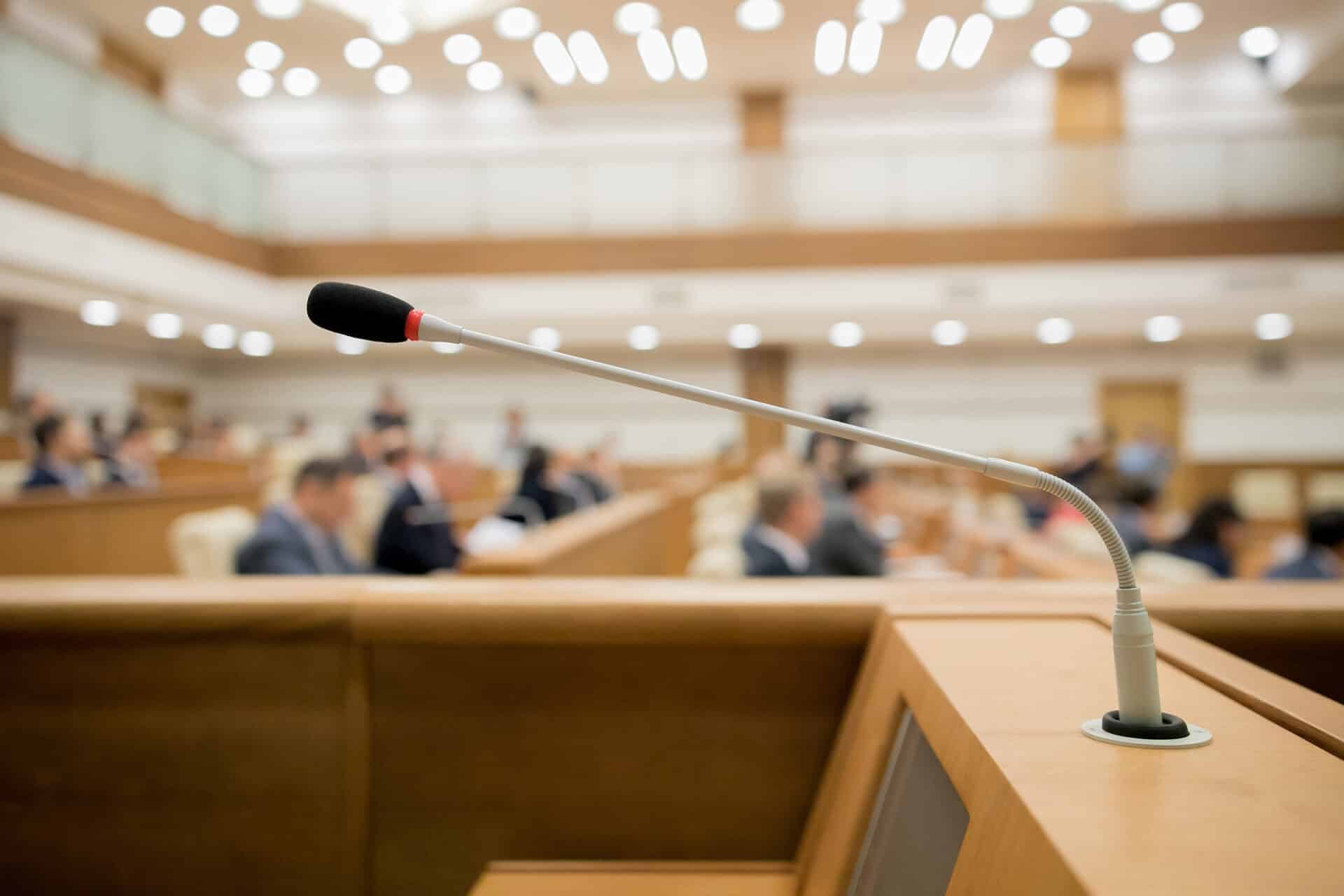 A microphone is prominently positioned at a podium in a large conference room. In the background, people in formal attire are seated in rows, listening attentively. The room has bright lighting and a modern design.