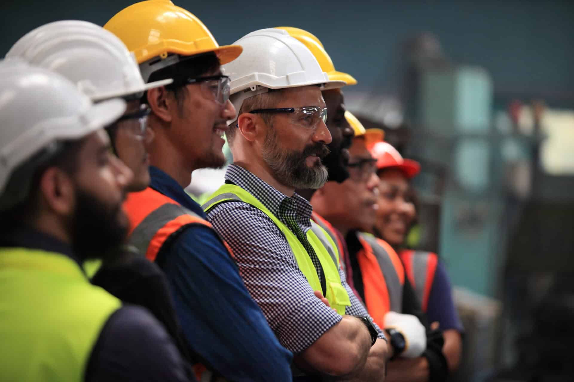 A group of construction workers wearing safety vests, helmets, and glasses stand together, smiling and looking forward. They are inside an industrial setting. The group includes men with diverse beards and mustaches.