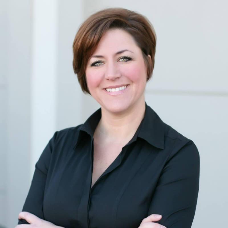 A person with short brown hair is smiling and standing with arms crossed. They are wearing a black shirt, in front of a plain, light-colored background.