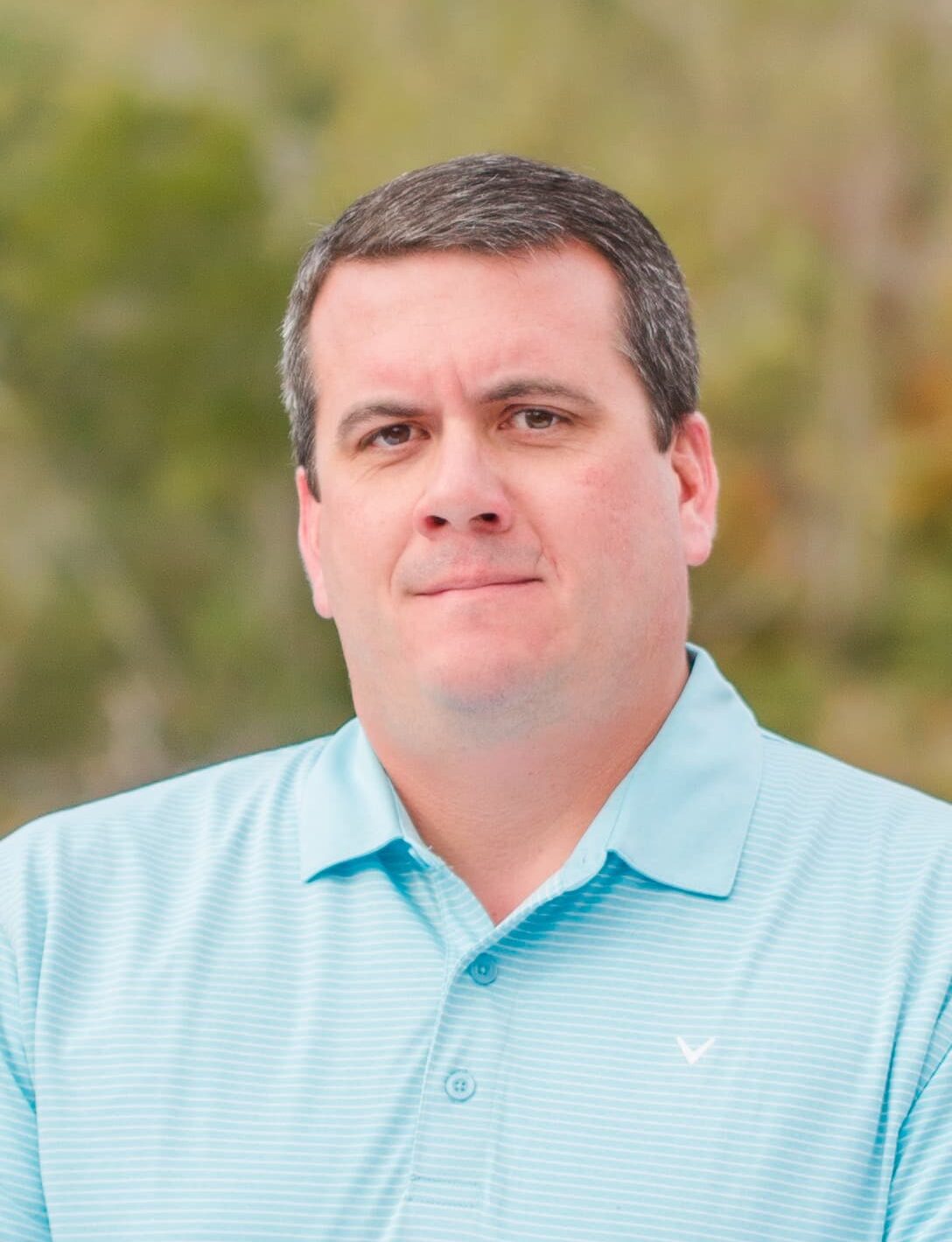 A man with short dark hair is wearing a light blue polo shirt. He stands against a blurred outdoor background with green foliage.