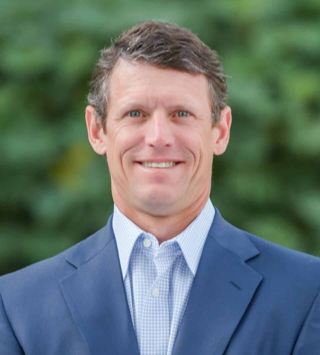 A man in a navy blue suit and light blue checkered shirt is smiling at the camera. The background is blurred greenery.