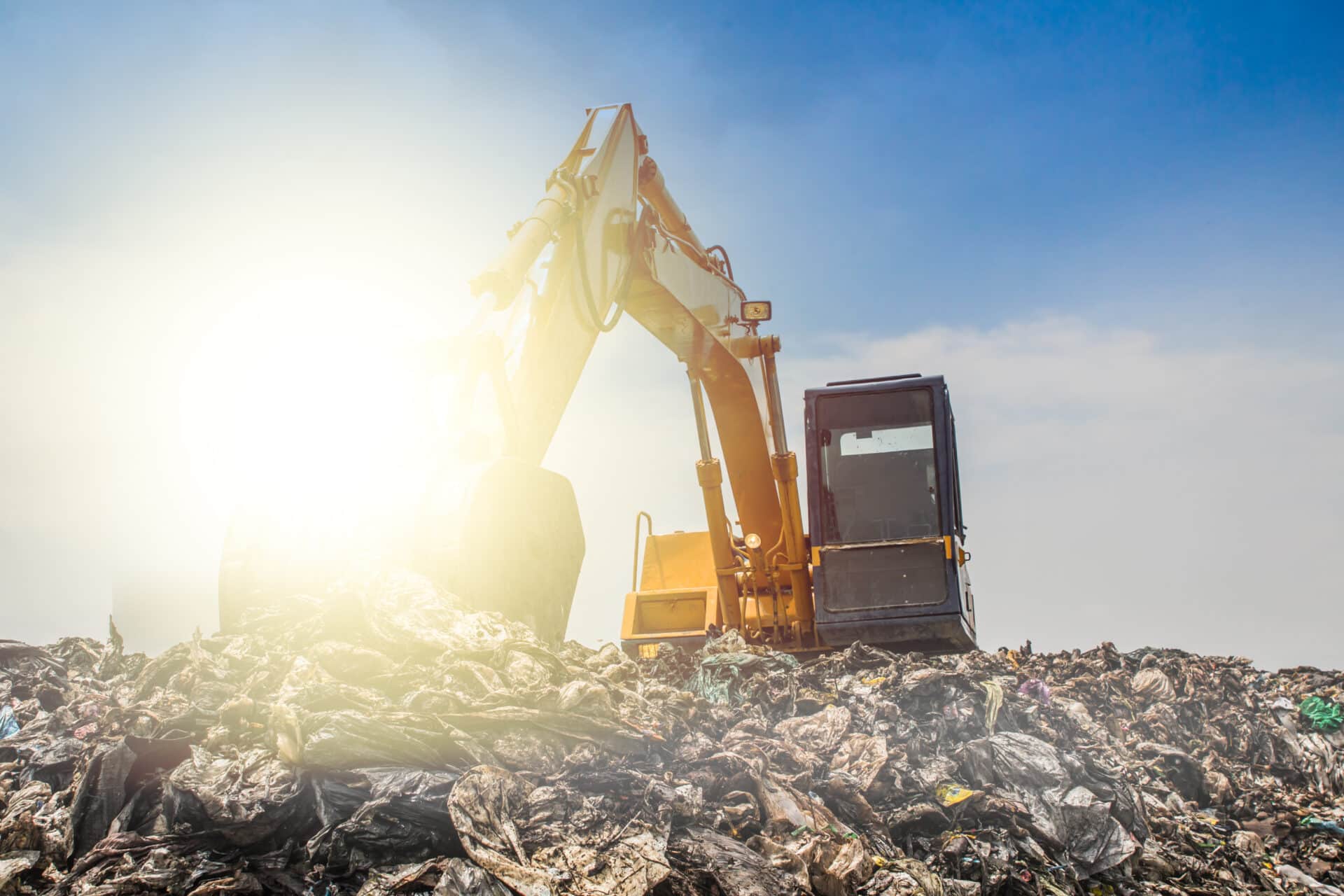 A yellow excavator sits atop a large pile of waste under a clear blue sky. The sun shines brightly, casting light onto the scene, highlighting the machine and the surrounding debris.