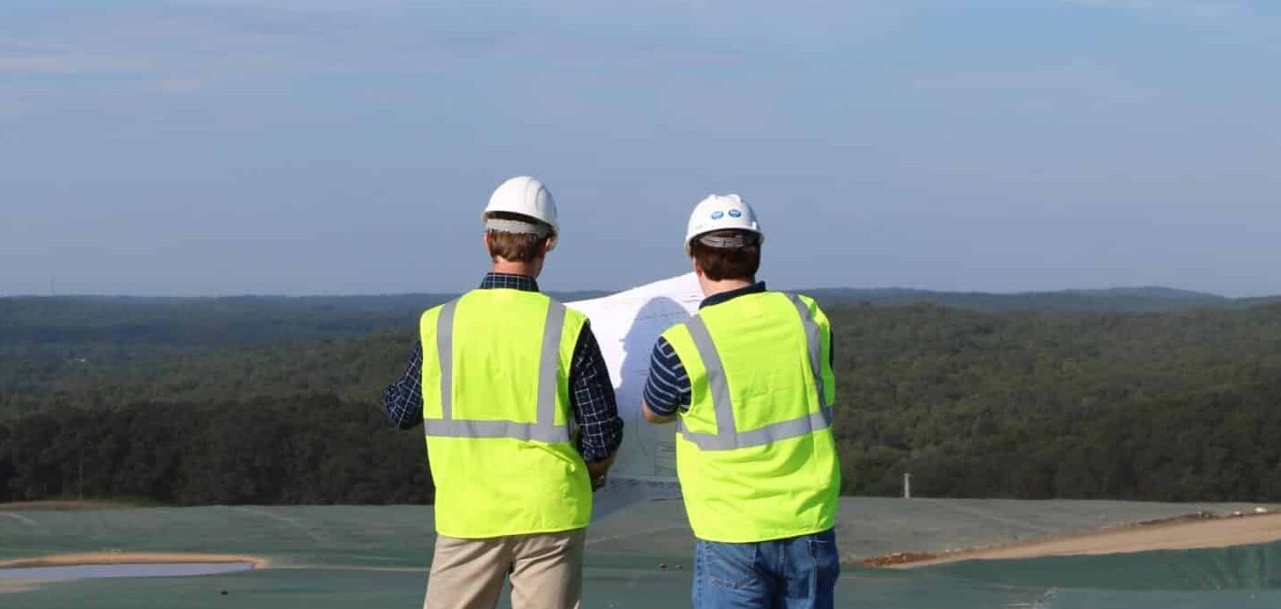 Two construction workers wearing hard hats and reflective vests are standing on a hill, looking at plans. They overlook a vast landscape with trees under a blue sky.