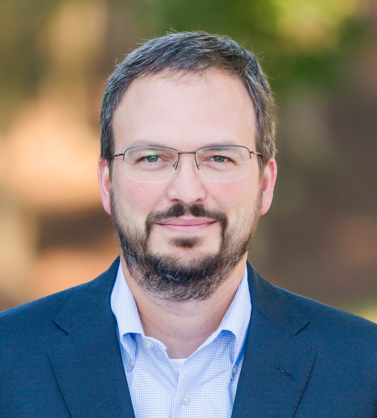 A man with short dark hair and a beard wearing glasses, a blue suit jacket, and a light blue collared shirt. The background is blurred with greenery and warm tones.