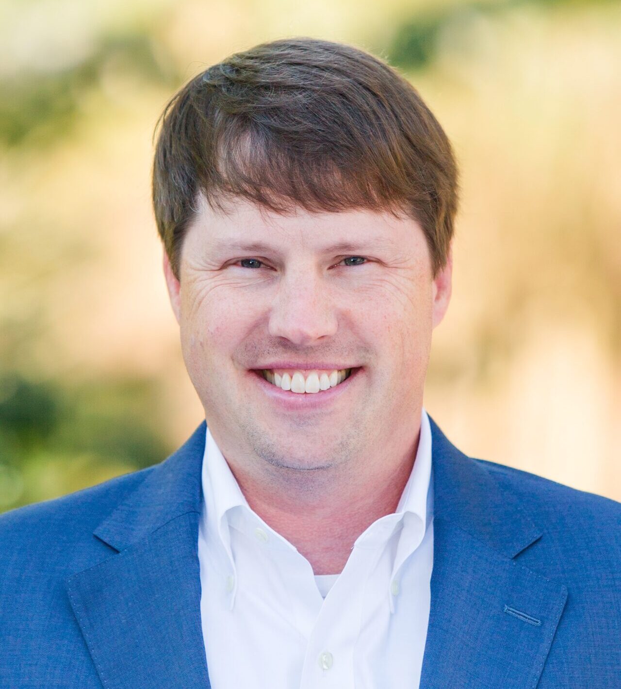 A man with short brown hair is smiling at the camera. He is wearing a blue suit jacket over a white shirt. The background is blurred with green and beige tones.