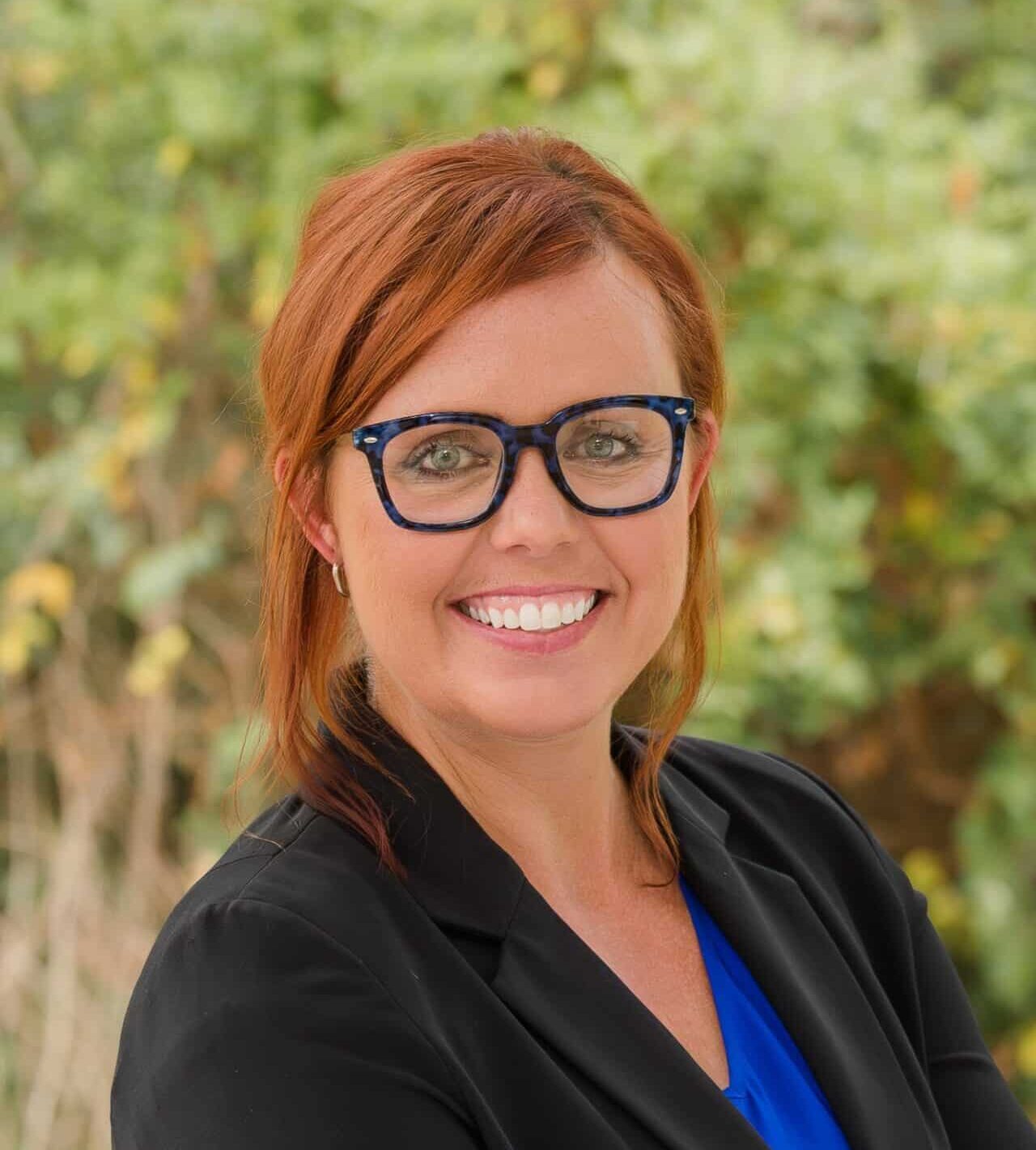 A person with auburn hair and glasses smiles, wearing a black blazer over a blue top. The background features green foliage.