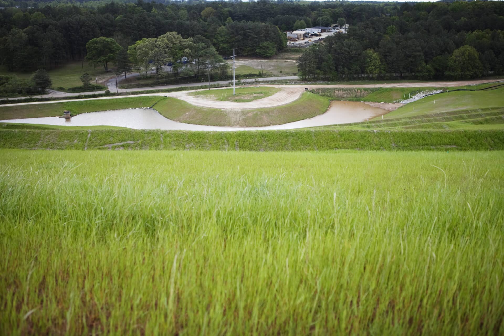 A grassy hillside overlooks a winding path and small ponds, surrounded by dense forest and trees under a cloudy sky. A few buildings are visible in the distance.