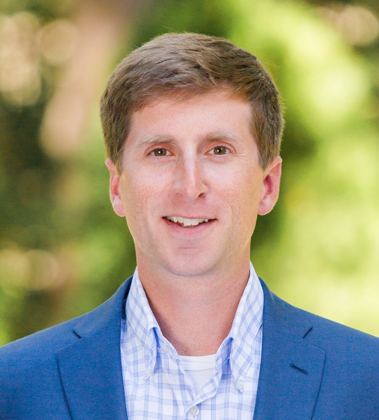 A person with short brown hair wearing a blue suit jacket and a checkered shirt, smiling against a blurred green outdoor background.