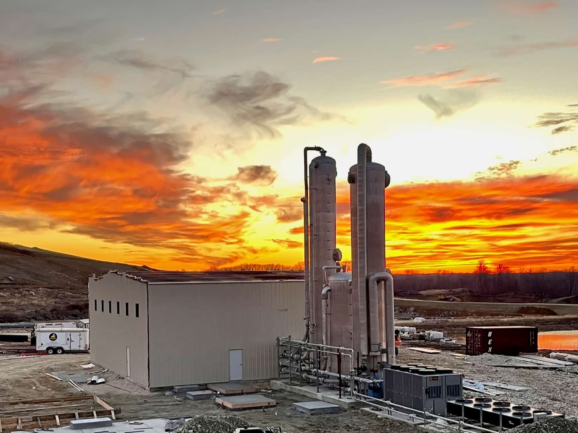 Industrial facility with tall towers stands against a vibrant sunset sky, featuring fiery orange and red clouds. The surrounding area includes open land, construction materials, and vehicles.