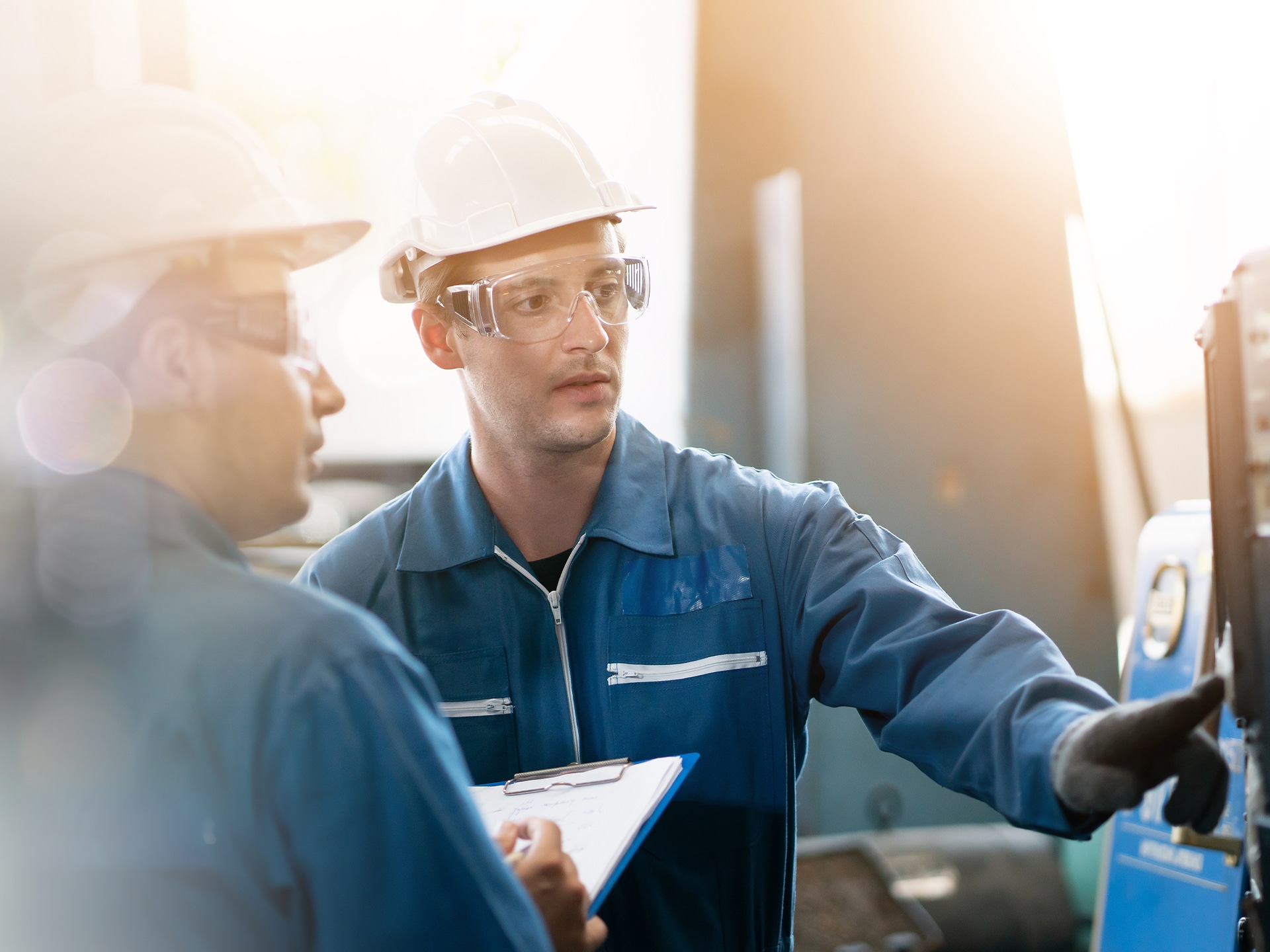 Two engineers in safety gear discuss at a worksite. They both wear hard hats and protective glasses. One holds a clipboard while the other points to machinery. The setting is industrial with bright lighting.