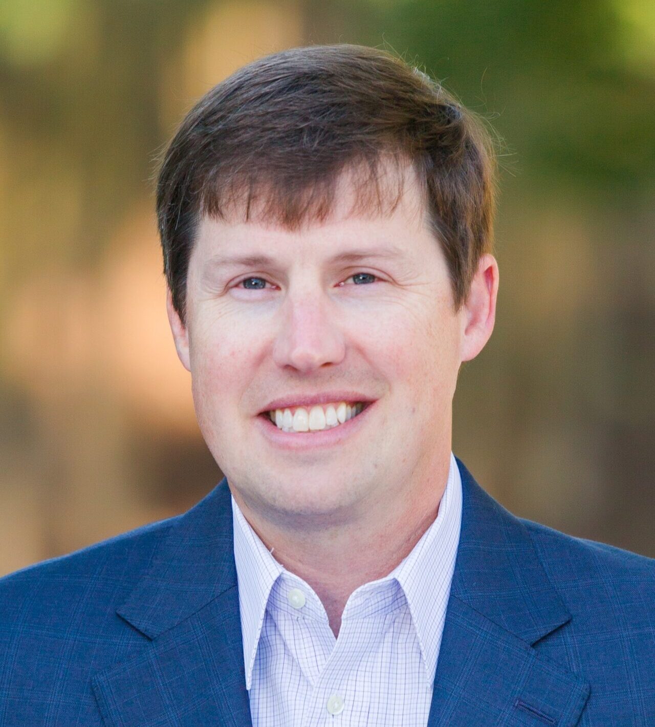 A man with short brown hair is smiling at the camera. He is wearing a blue blazer over a light checkered shirt. The background is blurred and features warm tones, suggesting an outdoor setting.