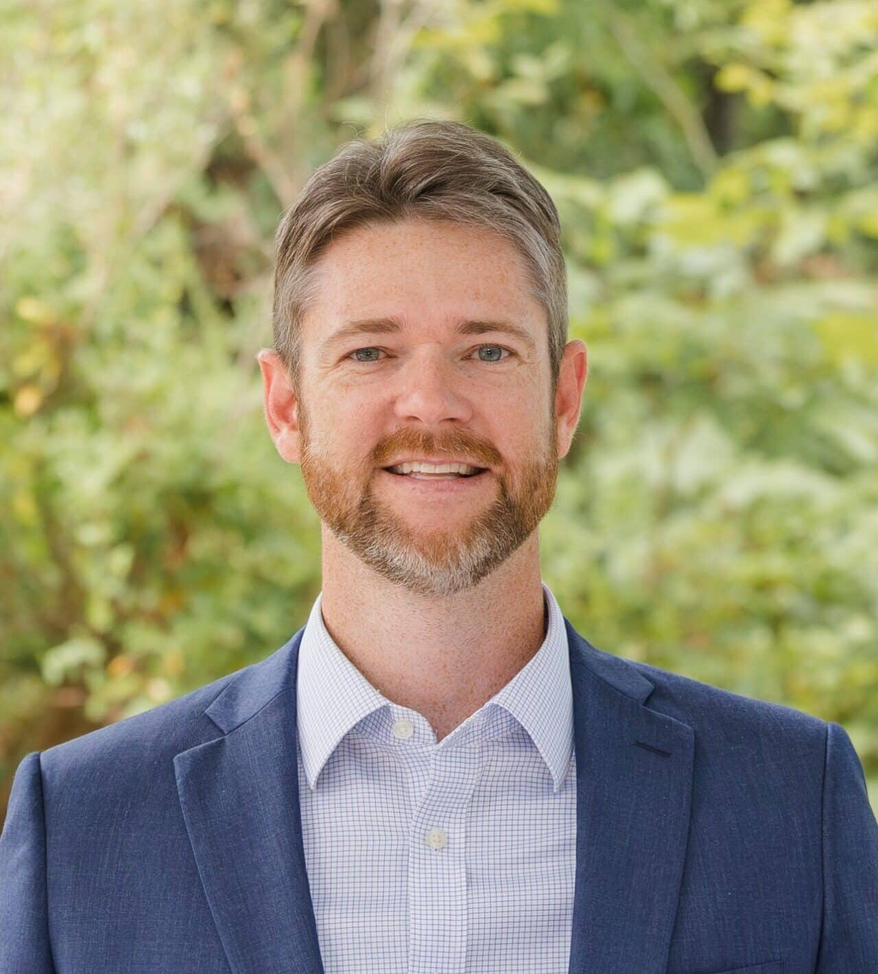 A man with short hair and a beard is smiling while wearing a blue suit jacket and a checkered shirt. He stands outdoors, with a blurred background of green foliage.