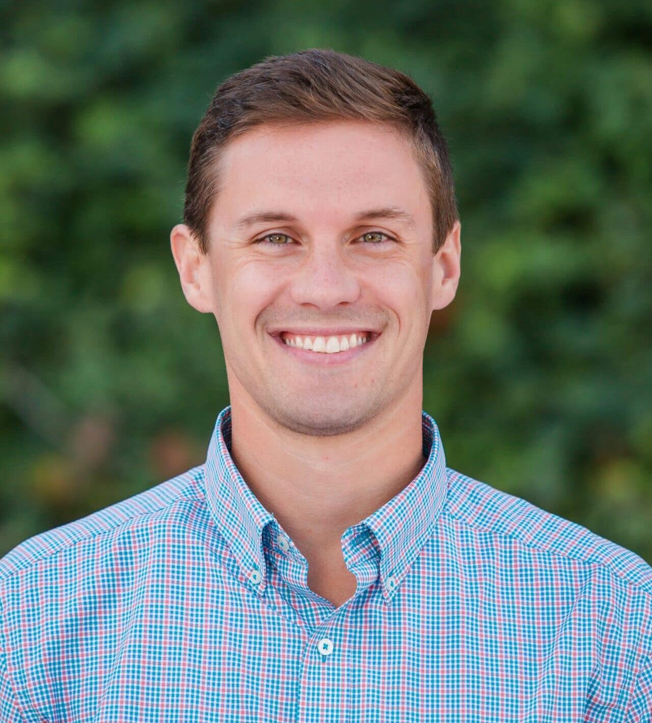 A person with short hair is smiling, wearing a checkered shirt, against a green, blurred leafy background.