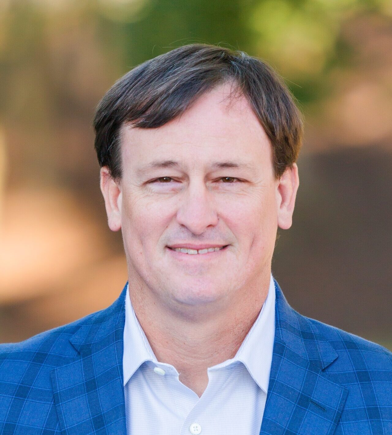 A man in a blue checkered blazer and white shirt is smiling at the camera. The background is blurred with warm, earthy tones, suggesting an outdoor setting.