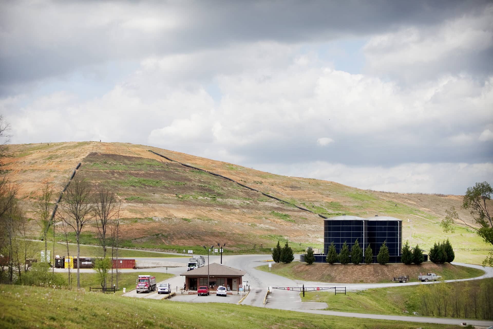 A landscape scene featuring a grassy hill with a few scattered trees under a cloudy sky. In the foreground, there is a small building, a black cylindrical structure, and some parked vehicles on a paved road.